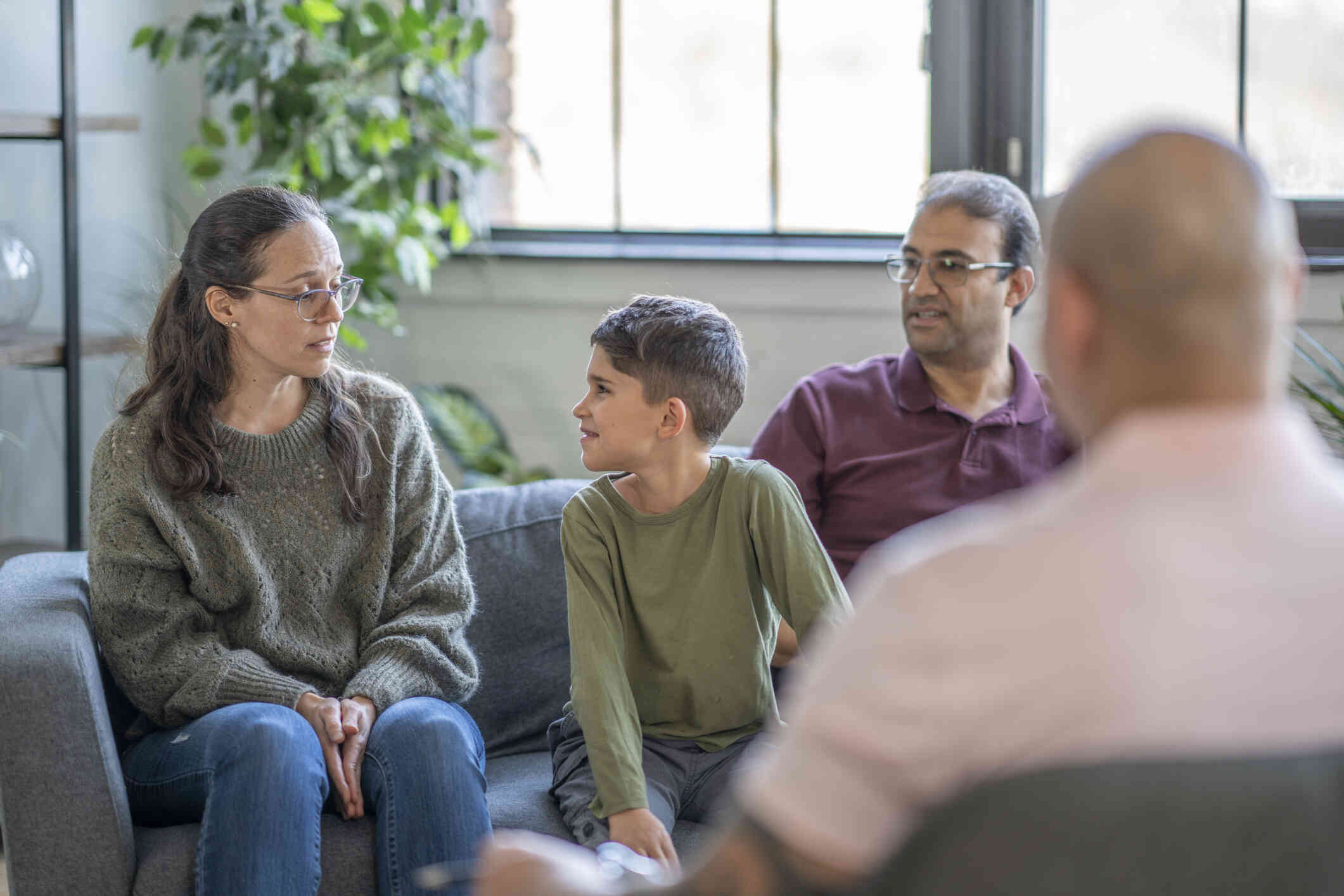 A mother, father, and young boy sit on a couch across from a therapist during a therapy session.